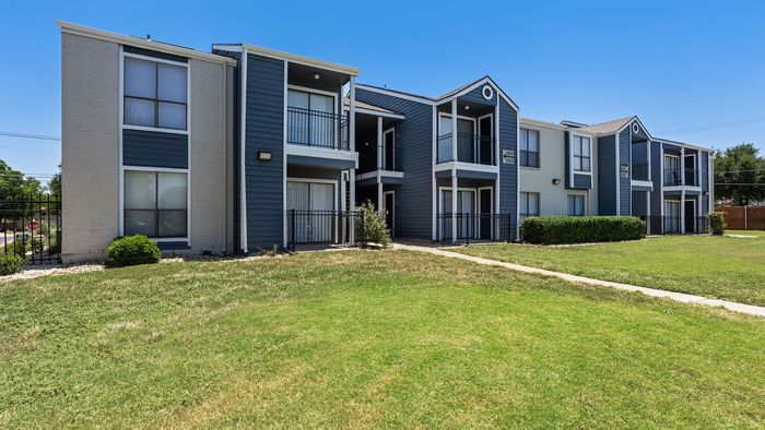 Charming apartment complex with modern blue and white exterior, offering spacious balconies and lush green surroundings under a clear blue sky.