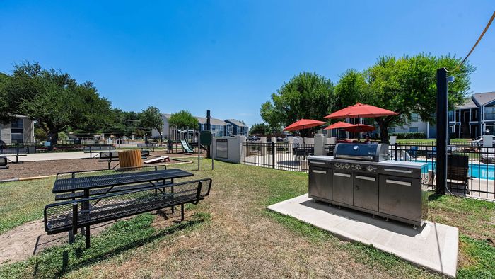 Sunlit outdoor space featuring modern grill, picnic areas, playground, and sparkling pool with red umbrellas.
