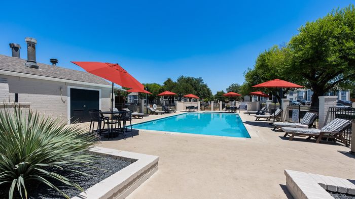 Inviting outdoor pool area with red umbrellas, comfortable lounge chairs, and lush greenery under a bright blue sky.