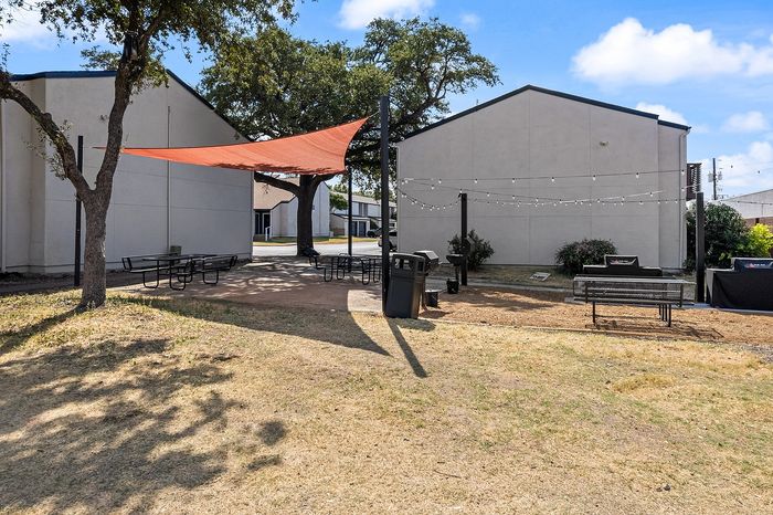 Charming outdoor communal space with shaded picnic tables, string lights, and a barbecue area set amidst lush greenery.