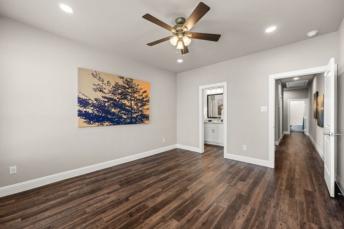 Elegant apartment interior showcasing a stylish room with rich wooden floors, modern ceiling fan, and a vibrant landscape painting, leading to a well-lit hallway.