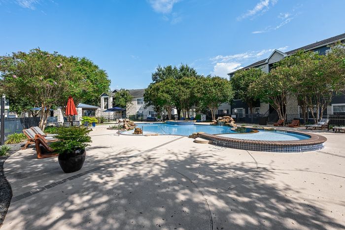 Luxurious apartment complex featuring a beautifully landscaped pool area with relaxing lounge chairs and vibrant greenery under a clear blue sky.