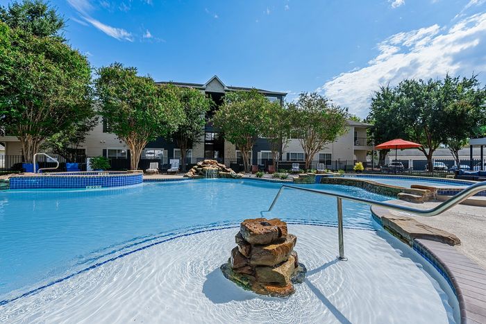 Luxurious apartment pool area featuring crystal-clear water, lush greenery, and inviting lounging chairs under a pristine blue sky.