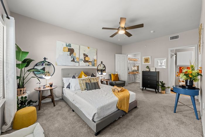 Modern bedroom with elegant decor featuring a plush queen bed, stylish side tables, vibrant yellow accents, and a cozy seating area. Includes spacious walk-in closet and contemporary ceiling fan.