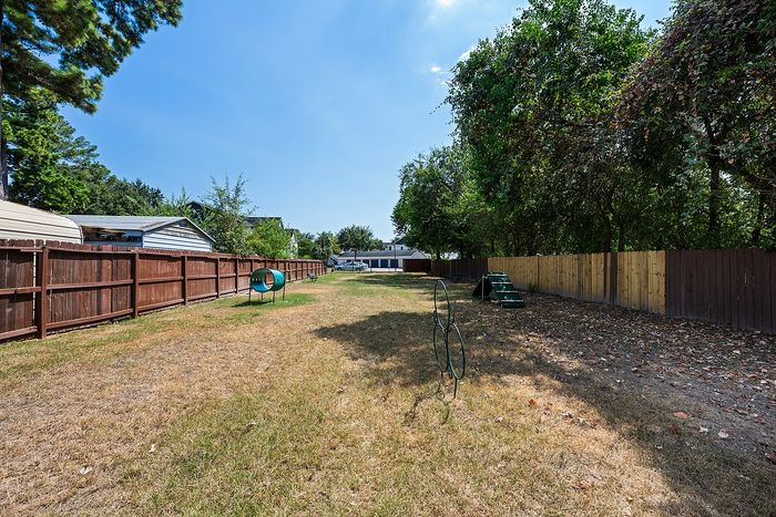 Spacious fenced backyard with lush greenery featuring a fun dog agility course under a bright blue sky.