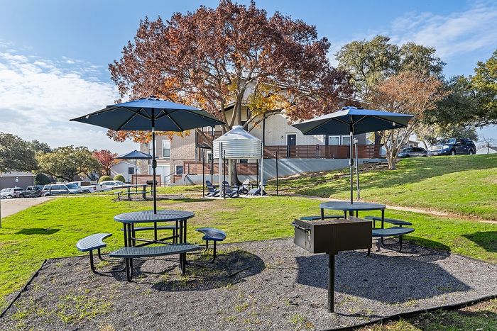 Charming outdoor community area with shaded picnic tables and barbecue grills, surrounded by lush greenery and scenic trees.