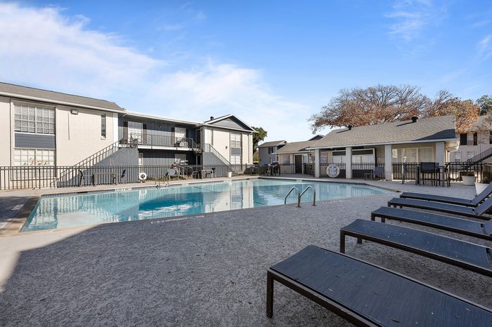 Inviting apartment complex with a pristine swimming pool, surrounded by modern architecture and sun loungers, set under a clear blue sky.
