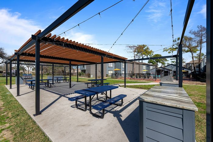Modern outdoor patio with stylish pergola, blue picnic tables, and barbecue area in a serene apartment community setting.