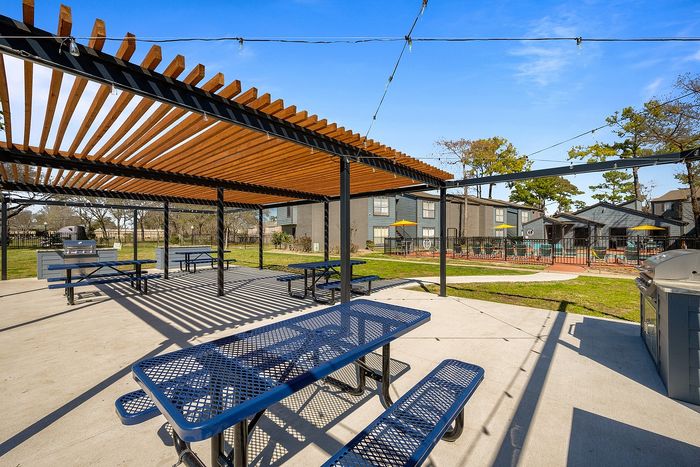 Charming outdoor picnic area with modern pergola and blue benches, perfect for gatherings, with a vibrant backdrop of a serene apartment community and lush greenery.