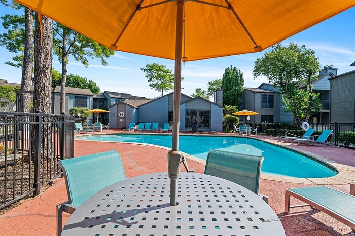 Inviting apartment pool area with relaxing lounge chairs, vibrant umbrellas, and lush trees, perfect for a sunny retreat.