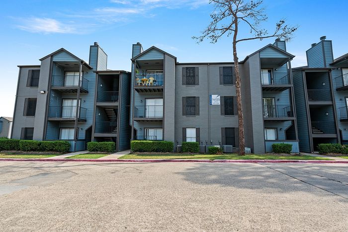 Modern three-story apartment building with balconies, surrounded by lush greenery, under a clear blue sky.