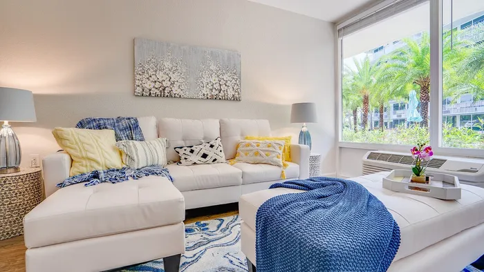 Chic living room with a plush white sofa, accented by colorful cushions and a blue throw, adjacent to a large window offering a scenic view of palm trees.