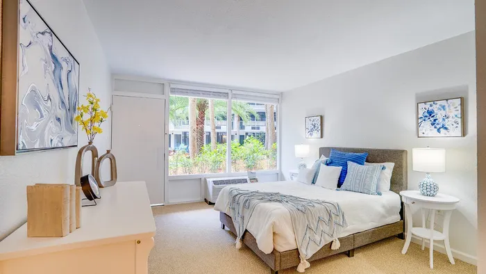 Bright and inviting bedroom with large windows, featuring a stylish bed adorned with blue and white accents, modern art, and vibrant decor.