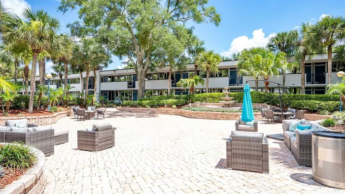 Charming outdoor patio with modern wicker furniture, lush palm trees, and a serene water fountain, set against a backdrop of elegant two-story apartments.
