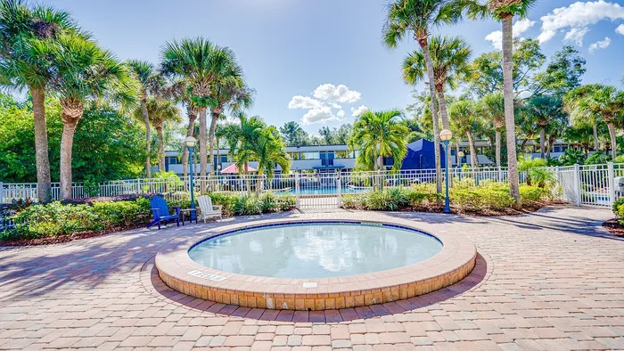 Vibrant outdoor scene with a round splash pool surrounded by tropical palm trees and comfortable seating, adjacent to a serene apartment complex.