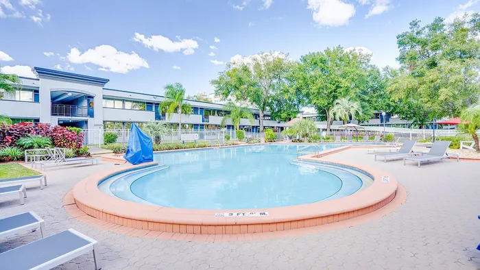 Inviting apartment complex with a sparkling pool, surrounded by lush greenery and modern lounge chairs under clear blue skies.