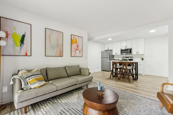 Modern open-concept living room with a stylish gray sofa, vibrant abstract art, and a sleek kitchen with stainless steel appliances.