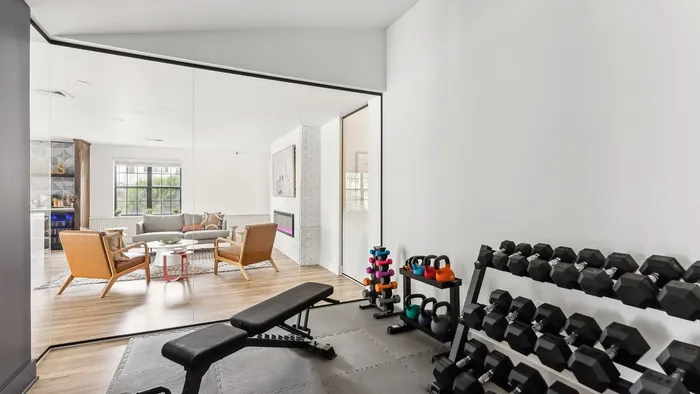 Modern apartment featuring a sleek home gym with weights and bench, opening into a bright, stylish living room with cozy seating and large windows.