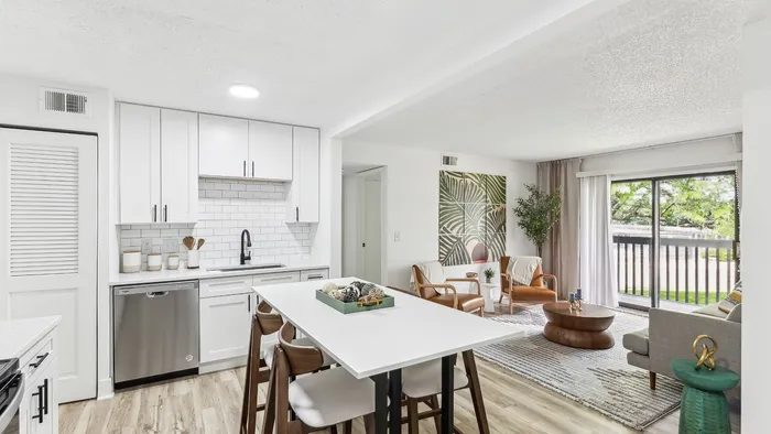 Bright, modern apartment interior showcasing an elegant, white kitchen with sleek cabinetry, an inviting open-plan living area with stylish decor, and a lush view from a large window, perfect for contemporary living.