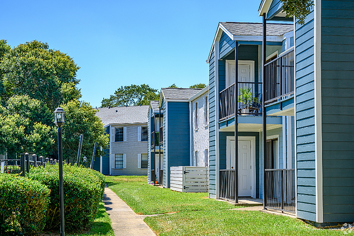 Charming apartment complex exterior with blue-gray siding, featuring private balconies and lush green landscaping under a clear blue sky.