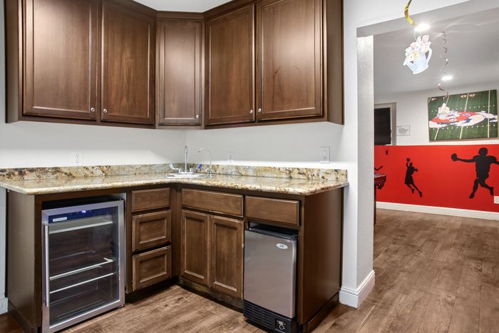 Chic corner kitchen with rich wooden cabinets, sleek granite countertops, and a modern mini wine fridge, opening to a vibrant sports-themed room.