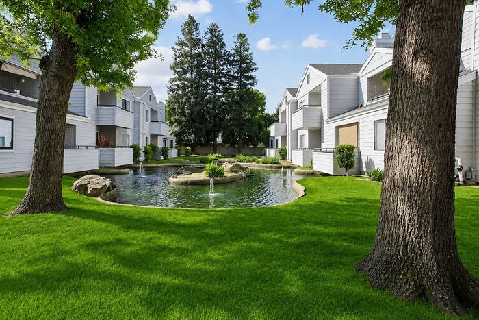 Charming apartment complex with lush green lawns and a picturesque pond, surrounded by modern white buildings under a bright blue sky.