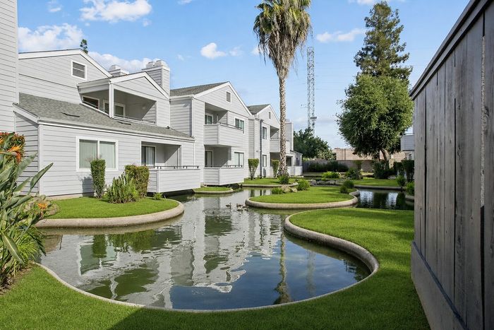 Modern apartment complex featuring elegant white facades surrounded by lush greenery and a serene water feature.