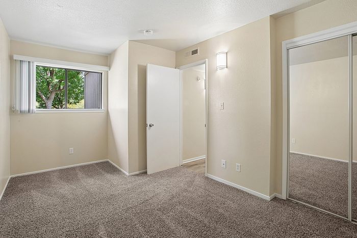 Bright, cozy bedroom with plush carpeting, mirrored closet doors, and a large window offering a view of lush greenery.