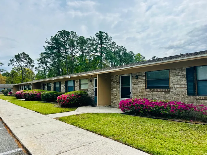 Charming brick apartment with lush greenery and vibrant flower bushes, nestled in a serene wooded area.
