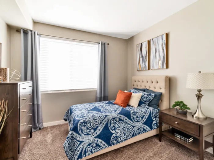 Dog-Friendly Townhomes in St. George, UT - Grayhawk Apartments at River's Edge - Cozy Bedroom With Tufted Headboard, Large Window With Blinds And Curtains, Nightstand Lamp, And Soft Carpet.