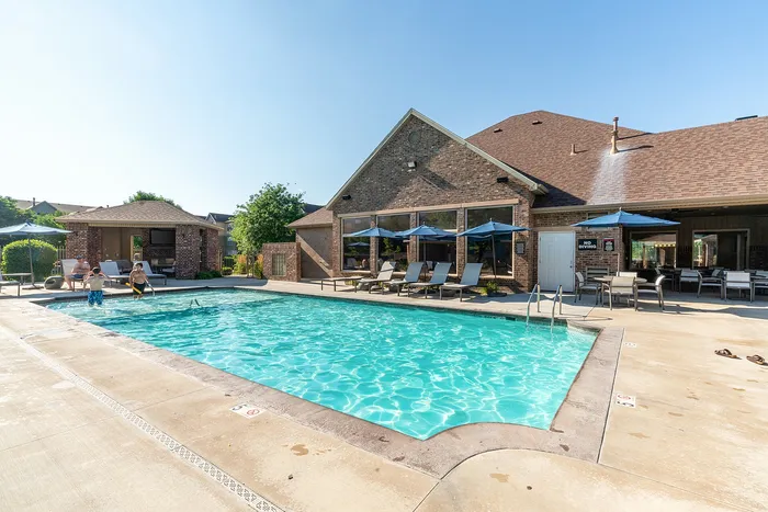 Enjoy a sunny day at this inviting apartment complex poolside with cozy loungers, shaded tables, and a charming brick clubhouse.