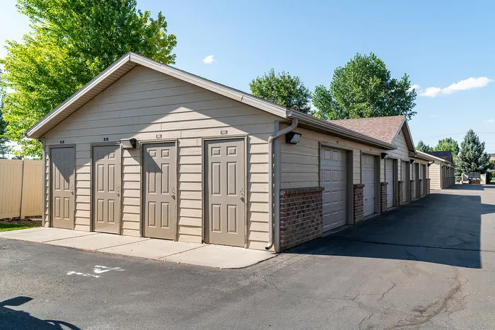Charming outdoor view of spacious individual storage units with beige doors and red brick accents, surrounded by lush greenery and clear blue skies.