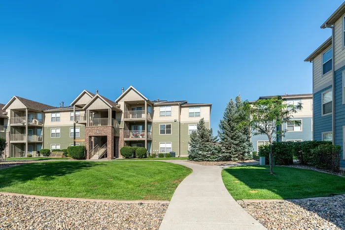 Charming apartment complex featuring three-story buildings with balconies, surrounded by lush green lawns and walking paths under a clear blue sky.