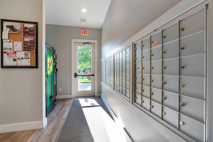 Bright and welcoming apartment entrance featuring secure mailboxes, a bulletin board with community notices, and a modern vending machine.