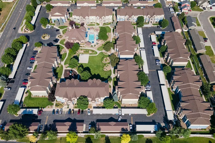 Aerial view of a picturesque apartment complex featuring lush green lawns, a sparkling swimming pool, and ample parking spaces surrounded by well-maintained residential buildings.