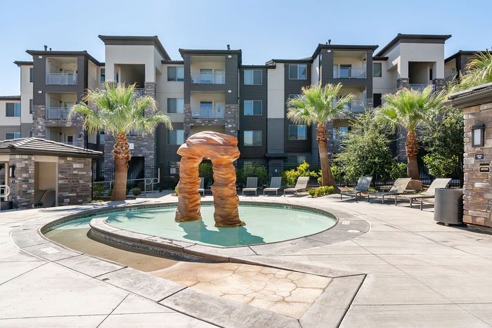 Luxurious apartment complex featuring a sparkling resort-style pool with stone arch structure, surrounded by lush palm trees and sleek lounge chairs.