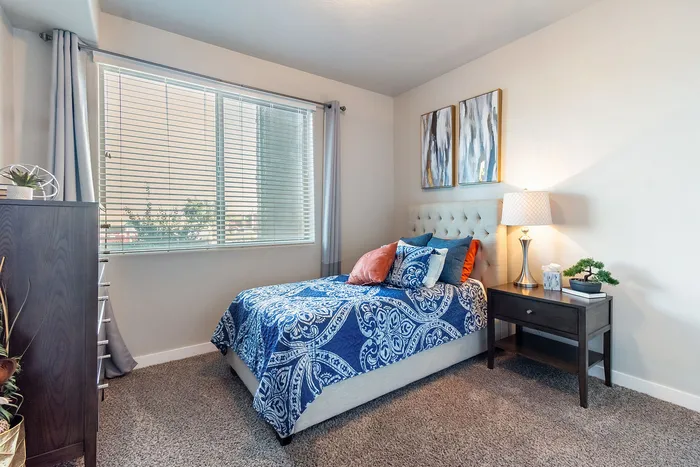 Dog-Friendly Townhomes in St. George, UT - Grayhawk Apartments at River's Edge - Cozy Bedroom With Tufted Headboard, Large Window With Blinds And Curtains, Nightstand Lamp, And Soft Carpet.