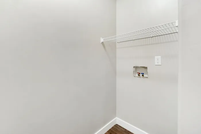 Bright utility room with sleek white walls, a mounted wire shelf, and convenient laundry hookups, perfect for modern functionality.