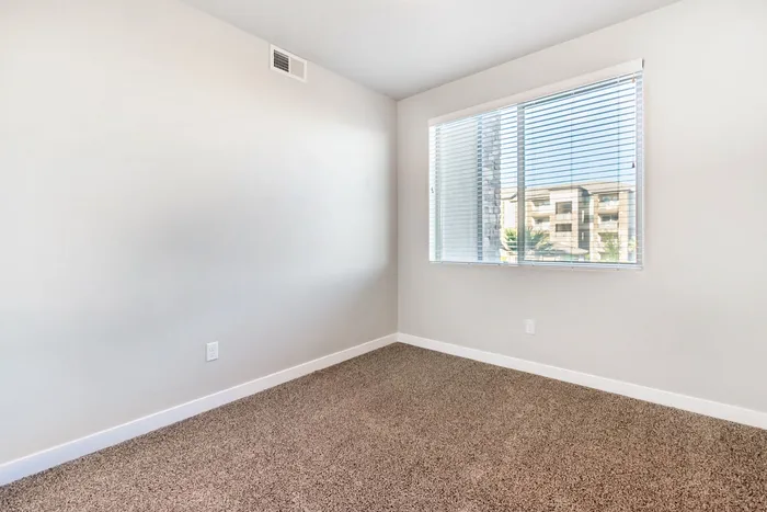 Bright empty room with plush carpeting, white walls, and large window with blinds, offering a view of neighboring buildings.