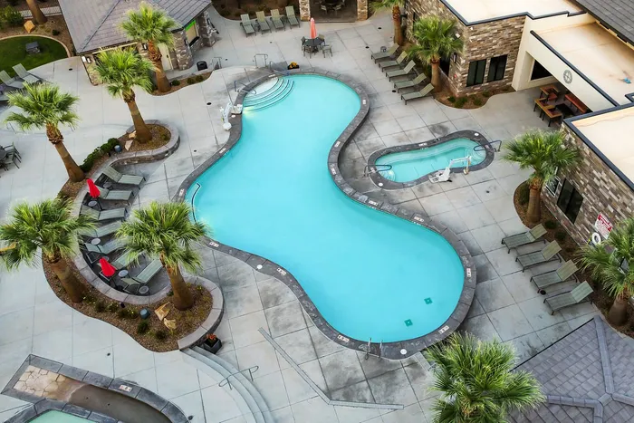 Luxurious apartment pool area with a uniquely shaped swimming pool, vibrant palm trees, and stylish poolside loungers under red umbrellas.