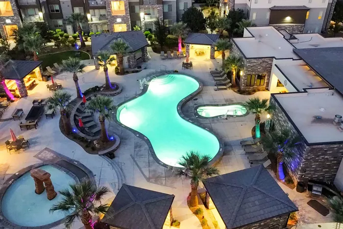 Luxurious apartment complex with a glowing resort-style pool, surrounded by palm trees and cozy cabanas, perfect for relaxing evenings.
