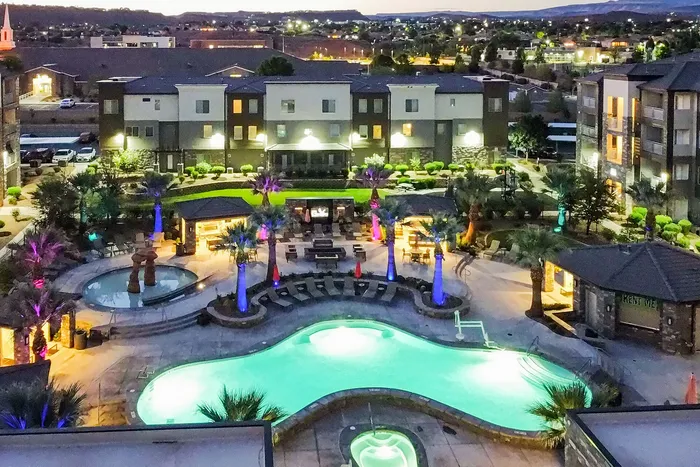 Vibrant apartment complex at night featuring a luxurious pool with colorful lighting, surrounded by lush greenery and modern buildings.