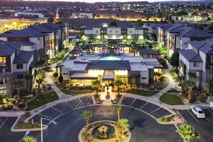 Stunning aerial view of a modern apartment complex featuring a central illuminated pool, lush landscaping, and surrounding buildings at dusk.