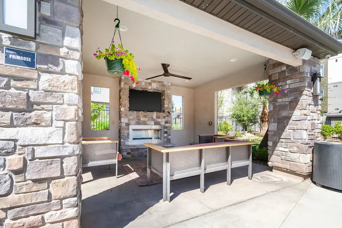 Charming outdoor lounge area with rustic stone fireplace, ceiling fan, and vibrant hanging plants, perfect for relaxation and entertaining.