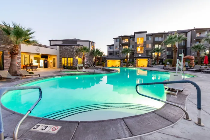 Luxurious apartment complex with an inviting, illuminated outdoor pool surrounded by palm trees and lounging areas.