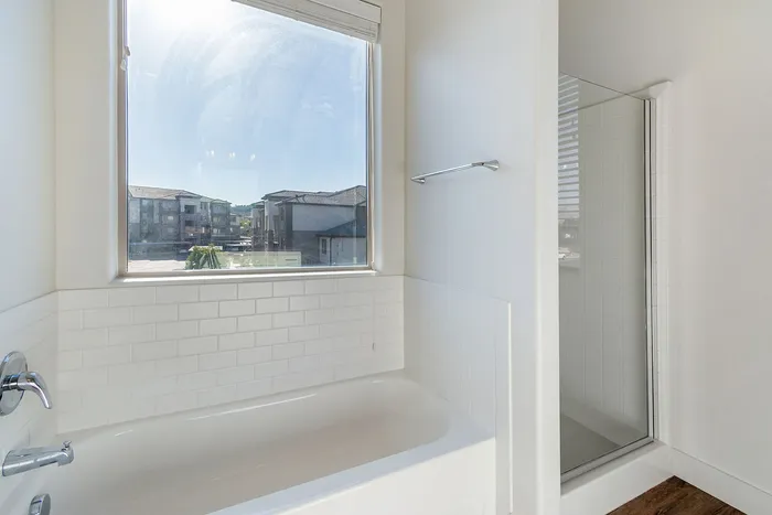 Bright bathroom with large window, featuring a luxurious bathtub and modern shower, offering serene views of the neighborhood.