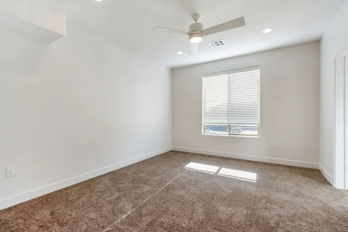 Inviting bedroom with plush carpeting, large window with blinds allowing natural light, and a modern ceiling fan for comfort and style.