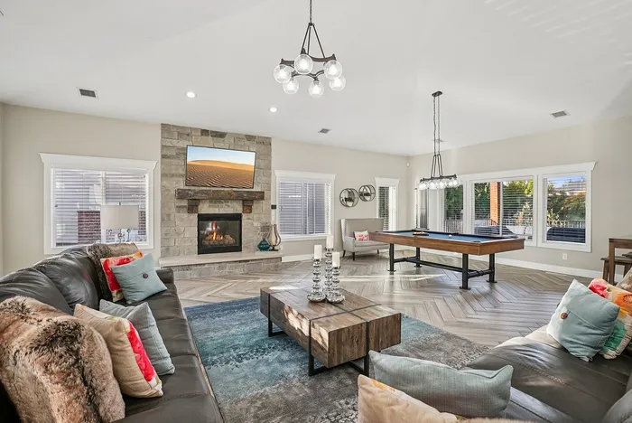 Contemporary living room with stylish leather sofas, vibrant cushions, a cozy stone fireplace, and an elegant chandelier. The room features a sleek pool table, large windows for natural light, and a modern rustic coffee table.