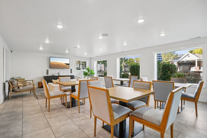 Bright and elegant community dining room with modern wooden furniture, large windows showcasing lush greenery, and a sleek mounted TV for an inviting atmosphere.