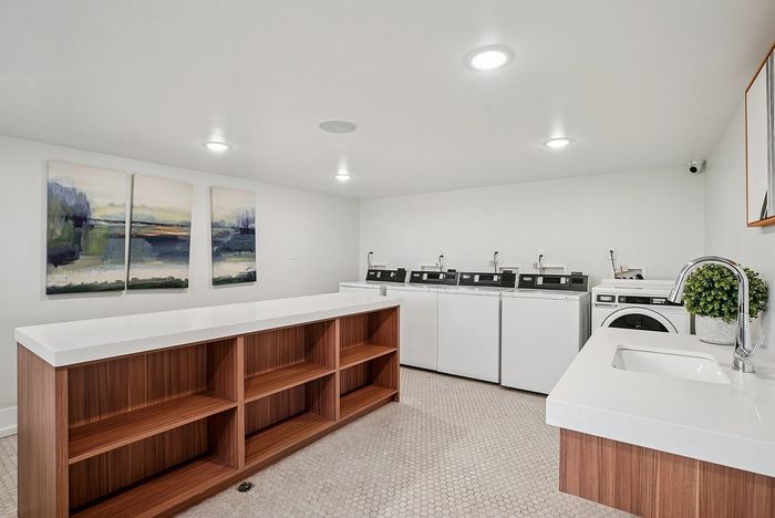 One-Bedroom Apartments in Ogden, UT - Pinebrook Apartments - Bright Laundry Room With Multiple Washers And Dryers, Folding Counter, Wood Cubbies, Wall Art, And White Tile Flooring.